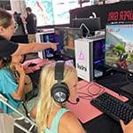 Two young girls are seated at desktop computers playing 'Minecraft' while a woman wearing a Super Girl Gamer Pro shirt assists them.